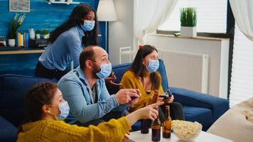 Sad friends because loosing playing games with wireless controller wearing face mask to prevent getting sick with corona in time of social pandemic. Group of people enjoying time together. photo