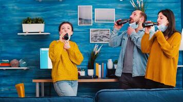 Group of friends toasting with bottles of beer wearing protective mask keeping social distancing against spreading covid 19 virus, enjoying time in living room. People socializing during outbreak photo