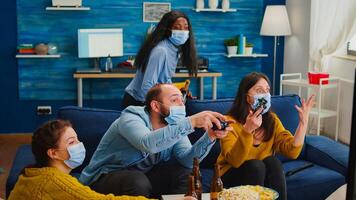 People screaming to each other loosing game playing at new normal party during social pandemic with face mask, keeping distancing against spreading covid 19 virus sitting on couch in living room photo