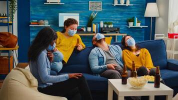 Group of multi ethnic friends watching comedy show on tv laughing wearing face mask to prevent infection with covid 19, during global pandemic having fun sitting on couch keeping social distance photo