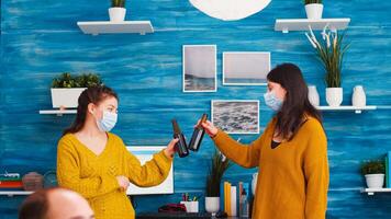 Joyfull women with protective mask discussing in living room respecting social distancing against spreading covid 19 virus. People socializing during outbreak clincking bottles of beer enjoying time photo