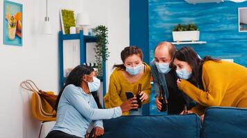 Group of multi ethnic friends hanging out in living room watching clip on smartphone sitting on couch respecting social distancing wearing face mask during global pandemic at new normal party photo