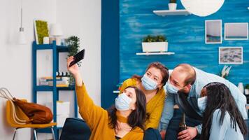 Multi-ethnnic friends with protective masks partying with beer taking selfie during covid 19 outbreak, new normal lifestyle concept with people having fun in living room respecting social distance photo