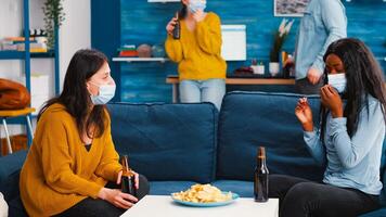 Mixed races women sitting on sofa discussing taking off protective mask eating snacks holding bottles of beer spending time at new normal party. Keeping social distancing against coronavirus pandemic photo