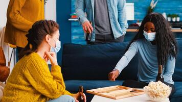 Cheerful woman using dice playing backgammon with multiethnic friends wearing face mask as prevention against covid 19 virus during global pandemic sitting on couch drinking beer and eating popcorn photo