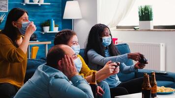 Excited funny young friends with masks holding controllers playing game at home during new normal party. Overjoyed man and woman winning videogame having fun together on sofa celebrating victory photo