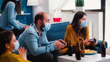 Excited diverse friends winning games using joystick having fun wearing face mask to prevent spreading of coronavirus in time of global outbreak. Gaming competition enjoying beer and popcorn. photo