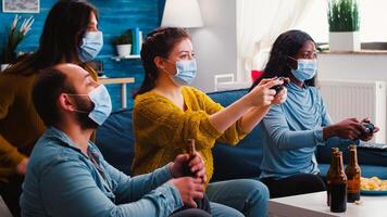 Excited multiracial gamer women sitting on couch playing games on console during new normal party wearing protective masks. Friends using wireless controllers keeping social distancing photo