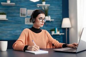 Student with headphones writing notes on notebook while searching communication information using laptop computer. Concentrated teenager making homework while sitting at desk in living room photo