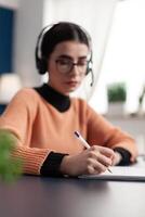 Young college student writing communication information on notebook for online school exam using e-learning platform. Student with headphone doing homework while sitting at desk table in living room photo