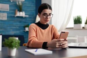 Student looking at smartphone screen while texting message sitting at desk table in living room. Student browsing lifestyle information on internet during coronavirus quarantine photo