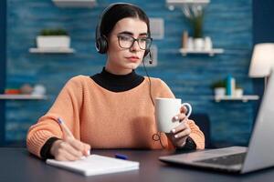 College student taking notes on paper while looking at university website searching information for online course. Youg woman using laptop computer while sitting at desk in living room photo