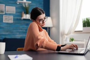 Teenager woman working at e-learning platform for university project during remote education. Student talking at phone about digital courses while sitting at desk in living room photo