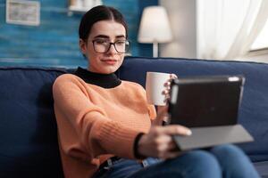 Student reading online book on digital application using tablet computer, sitting on sofa in living room. Teenager studying social communication using university mobile app drinking cup of coffee photo
