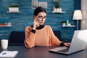 Student working at homework for college seminar using laptop computer browsing information on internet. Young woman talking on smartphone while sitting at table desk in living room photo