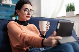 Student looking at online course about social media communication using mobile device computer, sitting on couch in living rom drinking cup of coffee. Teenager reading university communication online internet email photo