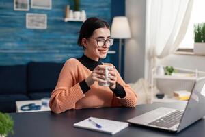 Smiling happy student holding cup of coffee in hands looking at laptop computer sitting at desk. Searching management advertising for university degree browsing e-learning online digital data photo