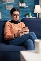 Student lying comfortable on sofa in living room while typing message to her freind using modern smartphone. Teenager chatting on social network looking at phone screen smiling photo