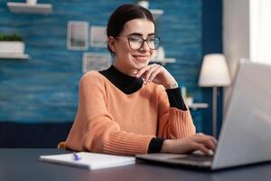 Student browsing on internet management information for university exam looking at e-learning platform. Woman doing her homework using laptop computer while sitting at desk table in living room photo