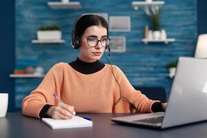 Student searching information on internet for university exam using laptop computer. Concentrated woman doing her homework using e-learning platform while sitting at desk in living room photo
