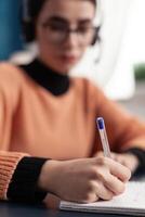 Selective focus on student hand taking notes on notebook for university exam before online meeting on zoom. Young woman with headphone doing homework while sitting at desk table in living room photo