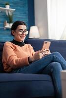 Smiling student relaxing on couch while talking with her friends on message about social media communication. Teenager woman chatting on social network using mobile app device photo