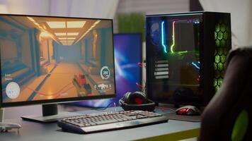 RGB powerful personal computer with first-person shooter game on screen in gaming studio with no people at desk. Empty cozy room with modern design is lit with warm and neon light. photo
