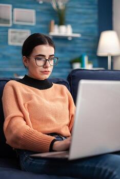 Student browsing online commerce information using laptop computer while sitting comfortable on sofa in living room. Woman studying marketing course using e-learning university platform photo