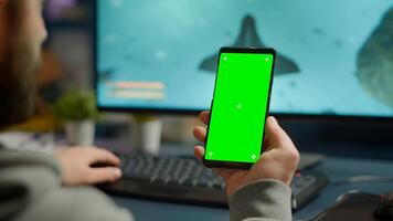 Competitive gamer looking at smartphone with green screen chroma key desktop playing online games on powerful computer during live tournament. Cyber player using phone with mock up isolated display photo