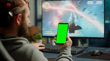 Concentrate gamer holding phone with green screen mock up chorma key display while playing games on powerful computer streaming online competition. Player using isolated desktop playing shooter games photo