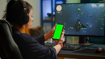 Professional cyber gamer looking at phone with greenscreen, chroma key, mock up desktop isolated display playing games on powerful computer. Player streaming shooter games wearing headset photo