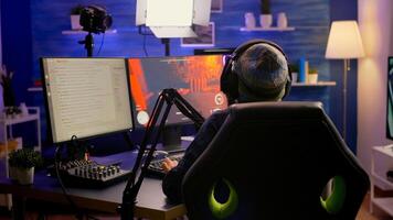 Back shot of pro streamer with headphone playing first person shooter game competition use RGB keypad. Player sitting on gaming chair using streaming equipment late at night in home studio photo