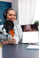 Woman smiling showing digital tablet with white screen. Blogger holding device with mockup template and isolated background while using microphone and headphones to stream podcast. photo