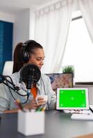 Woman vlogger showing digital tablet with horizontal green screen. Blogger holding gadget with chroma key and isolated mockup template while recording podcast with microphone and headphones. photo