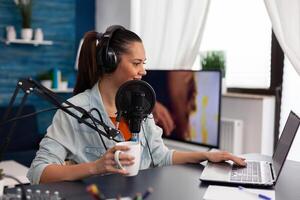Woman vlogger holding cup and streaming podcast using laptop. Influencer with headphones in home office recording livestream on computer. Blogger live broadcasting for social media with microphone. photo