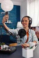 Woman influencer in home studio streaming live on smartphone. Content creator using microphone and mobile phone to stream and record for social media channel. Podcast professional home studio. photo