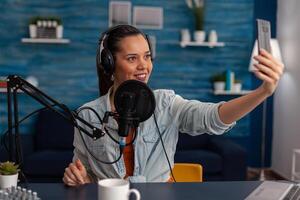 Woman vlogger with headphones streaming live on smartphone. Content creator using microphone and mobile phone to stream and record for social media channel. Podcast professional home studio. photo