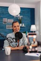 Smiling influencer streaming vlog in front of recording mobile phone. Content creator in home studio filming for social media. Woman broadcasting podcast with microphone and smartphone. photo