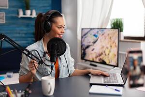 Woman in home studio recording podcast with mobile phone. Vlogger using headphones, microphone and laptop to stream online vlog. Content creator broadcasting with smartphone for social media. photo