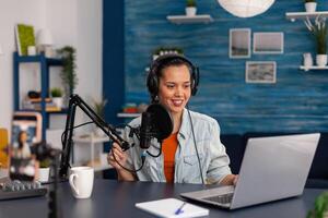 Social media influencer looking at laptop and recording . Woman vlogger streaming online podcast on smartphone. Content creator in home studio using audio broadcasting equipment. photo