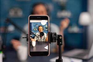 Closeup of mobile phone recording smiling woman vlogger on live stream podcast. Influencer using microphone and headphones, talking in front of phone camera and filming vlog for social media channel. photo