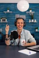 Portrait of happy woman facing camera recording in home studio. Vlogger with headphones creating content for online social media. Influencer streaming podcast using microphone and laptop. photo