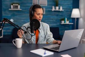 Woman vlogger streaming podcast and using laptop at desk. Influencer with headphones in home office recording livestream on computer. Blogger live broadcasting for social media with microphone. photo