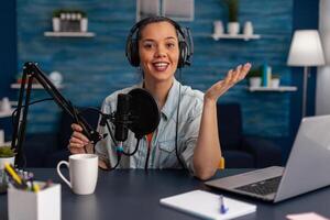 Portrait of woman smiling at camera in home recording studio. Vlogger with headphones creating content for online social media. Influencer streaming podcast using microphone and laptop. photo