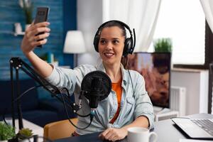 Woman content creator using headphones and microphone to stream live podcast on smartphone. Social media influencer recording online broadcast with mobile phone in home studio. photo