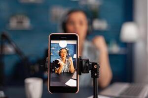 Closeup of smartphone recording woman influencer on live stream podcast. Vlogger talking in front of phone camera and filming for social media channel, using microphone and headphones. photo