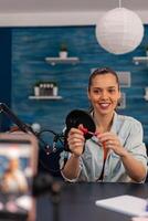 Smiling influencer filming lipstick review with mobile phone. Woman vlogger recording make-up recommendation with smartphone for social media channel. Online broadcasting in home studio. photo