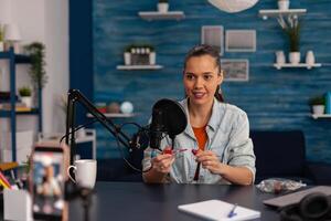 Woman vlogger recording product review of lipstick with smartphone. Smiling influencer filming make-up recommendation with mobile phone for social media channel. Online broadcasting in home studio. photo