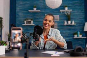 Smiling influencer recording mouse review with smartphone. Content creator in home studio with microphone and mobile phone doing product recommendation. Vlog podcast for social media. photo