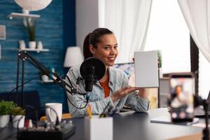 Smiling vlogger presenting white product box in front of camera. Woman content creator recording online vlog review for social media channel. Home recording studio with microphone and laptop. photo